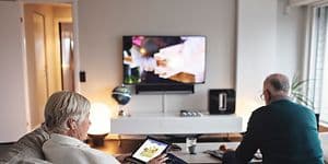 Eldre kvinne som sitter i sofa med nettbrett, mens eldre mann ser på TV i bakgrunnen